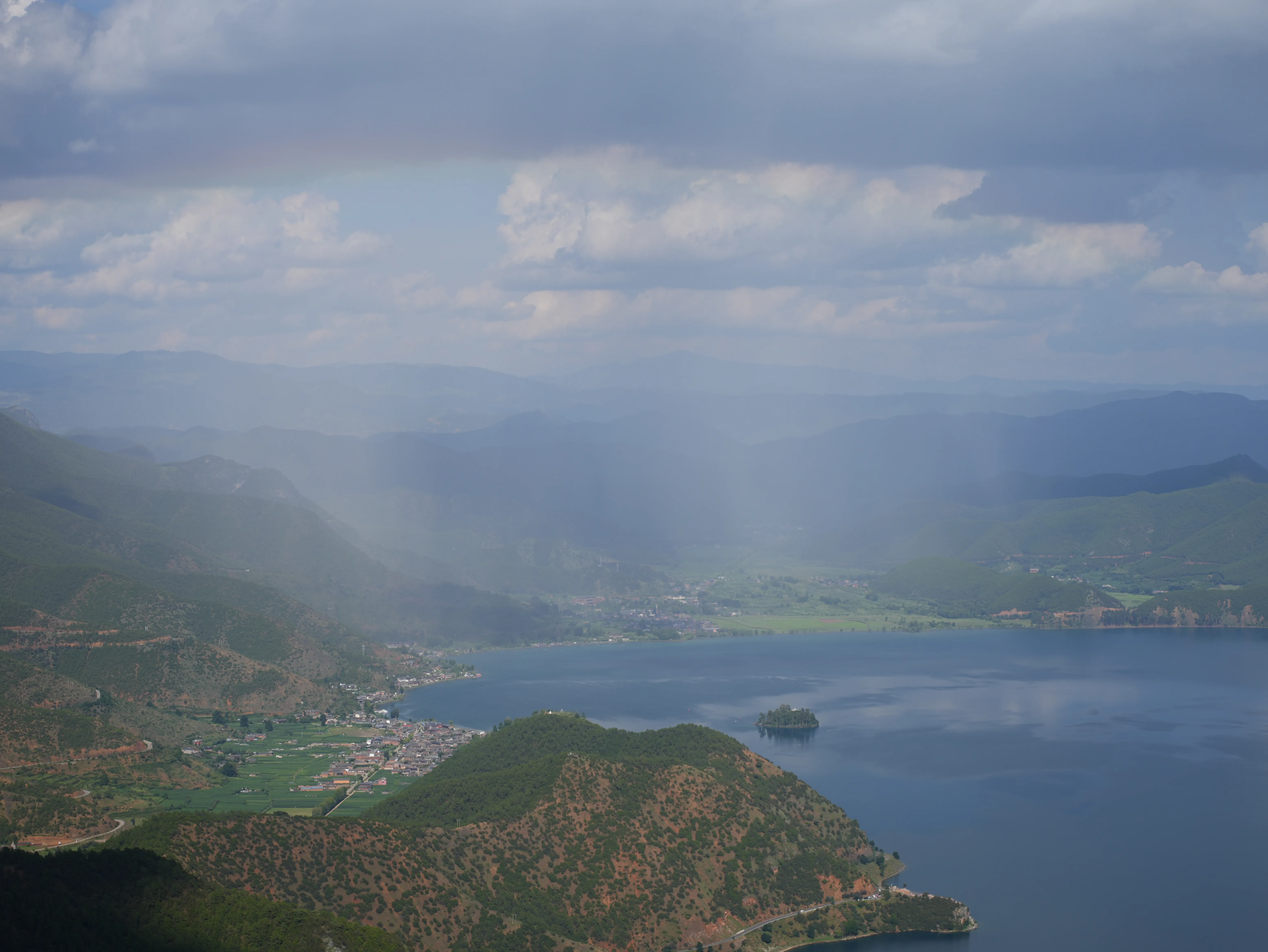 A mountaintop view overlooking Lugu Lake and the surrounding mountains, part of the landscape is shrouded in rain while a faint rainbow across the sky